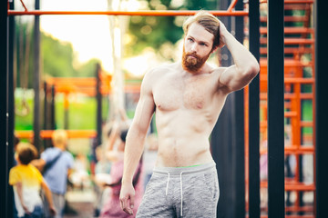 A handsome young Caucasian man with a naked muscular torso with long red hair and a beard posing on a street sports field, an outdoor gym. Sexy guy touches his hair