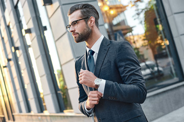 Naklejka premium Close up profile portrait of a successful young bearded guy in suit and glasses. So stylish and nerdy. Outdoors on a sunny street, fixing his cuffs