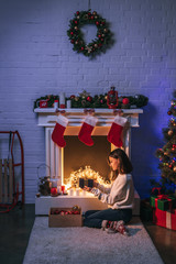 beautiful woman holding gift and sitting near christmas decorated fireplace and Christmas tree