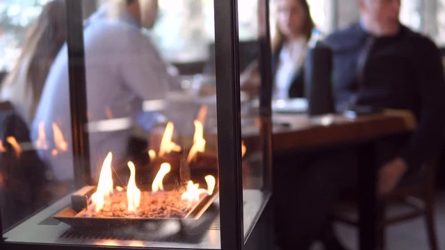 Stone gas fire pit with flames in modern fireplace in the restaurant with blurred people meeting at dining table in the background