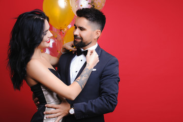 Side view of attractive lady in evening dress smiling and adjusting bow tie of handsome male while standing near bunch of balloons on red background. Smiling woman adjusting bow tie of man