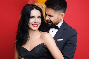 Charming young lady smiling and looking at camera while standing near bunch of balloons and crying guy in suit on red background. Cheerful woman and crying man near balloons