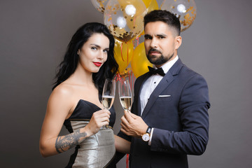 Attractive man and woman in elegant outfits holding glasses of champagne and looking at camera while standing near bunch of balloons on gray background. Elegant couple with champagne