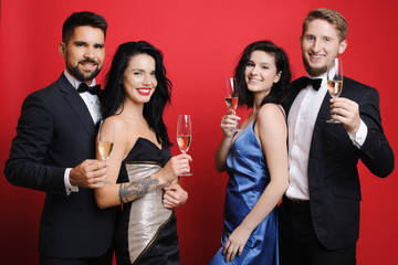  handsome men in elegant suits and charming ladies in evening dresses holding glasses of champagne and cheerfully smiling while standing on vibrant red background. Young couples posing with champagne