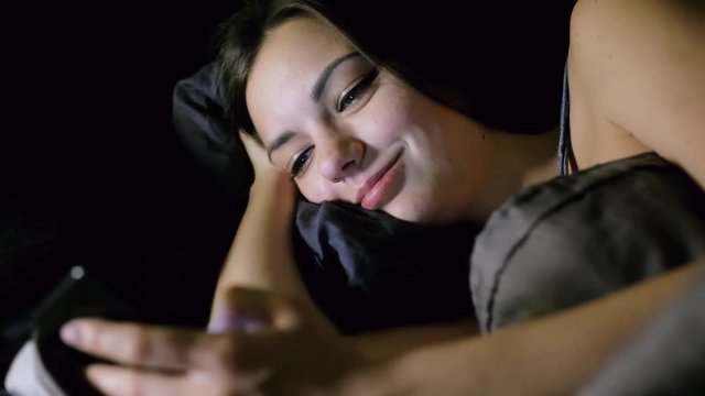 Girl surfing internet while lying in bed in the bedroom at night, looks photo, chating and smile