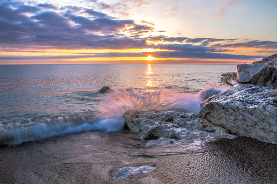 Waves Crashing On Beach. Wave crashing over a rocky coast with sunrise colors reflecting on the waters of Lake Huron In Lexington, Michigan.