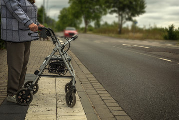 An old woman stands with her walking frame on a road and wants to cross it. Concept: mobility in...