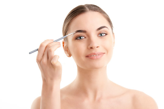 Studio shot of a beautiful young woman applying makeup to her eyebrow while standing at isolated white background. 