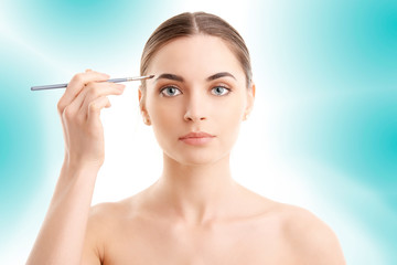 Studio shot of a beautiful young woman applying makeup to her eyebrow while standing at isolated light blue background. 
