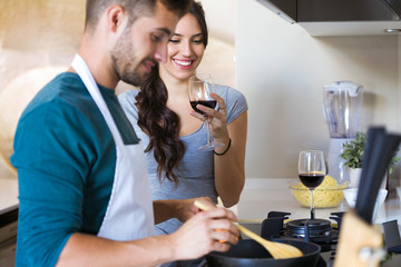Beautiful young smiling woman drinking red wine while her husband preparing lunch in the kitchen at home.