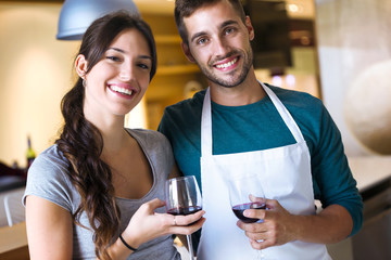 Happy young couple looking at camera while holding glasses of red wine in the kitchen at home.