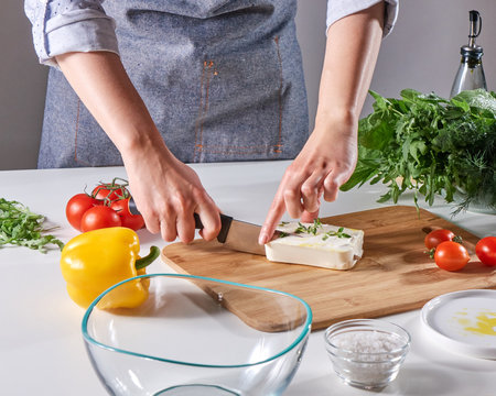 Woman's hands cut the cheese with the sprouts on the wooden board on the kitchen table with different vegetables. Step by step cooking healthy salad