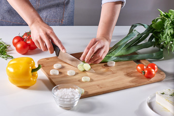 Fresh leek slices cut into the hands of a female cook on a wooden board on the kitchen table around a variety of organic vegetables. Cooking Healthy Salad