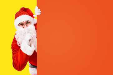 Fototapeta na wymiar Santa Claus with finger on lips asking for silence with colorful advertisement board and copy space