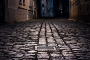 Leere Gasse, die nachts mit nassem Kopfsteinpflaster gepflastert ist, beleuchtet von Straßenlaternen mit einer Abwasserkappe im Fokus