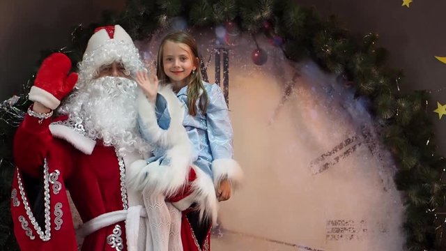 The joyful child and Father Frost welcome hand waves. Eve of a children's Christmas holiday. New Year's characters against the background of huge hours.