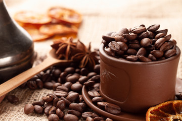 Wooden spoon with anise stars lies between the Turk for making Coffee and coffee, which are scattered near a brown clay Mug with a saucer and dried orange. A cup is overflowing with roasted coffee.