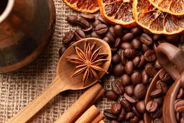 Wooden spoon with anise stars and cinnamon lie near the Turk for making Coffee, oranges, and overflowing Mug with coffee beans which are scattered on the table with burlap cloth on. Top view. Close up