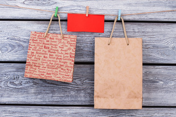 Gift bags hanging on rope with clothespins. Kraft paper shopping bags and red blank paper card on wooden background. Holiday sale concept.