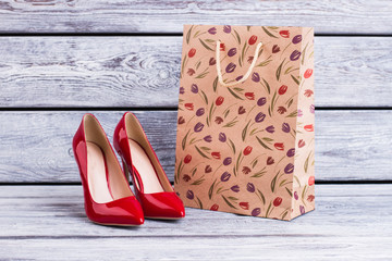 Red lacquered heels and shopping bag. Female leather stilettos and paper bag with spring tulip pattern. Female spring purchase.