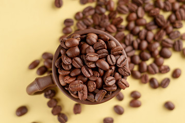 A brown earthenware mug overflowing with grains of fragrant aromatic coffee stands on a yellow cardboard background. View from above. Mug with coffee in the center and in focus.