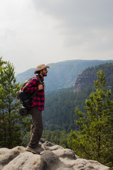 Young male hiker stand on the cliff in mountains with baclpack and hat