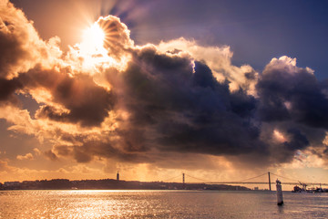 Landscape view on Lisbon ponte 25 de abril bridge, with sun ray and clouds in sky, boats and jesus christ statue