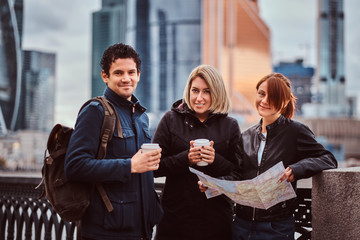 Group of smiling tourists holding the map and takeaway coffee in front of skyscrapers