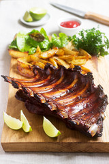 Homemade baked meat ribs served with french fries, herbs, lime and ketchup on a wooden board. Rustic style.