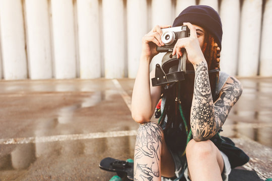 young girl with tattoos and dreadlocks in a blue hat photographs a vintage camera on the background of a concrete wall