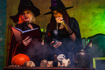 Image of smiling two witches in black hats reading book at table with pumpkin and skulls