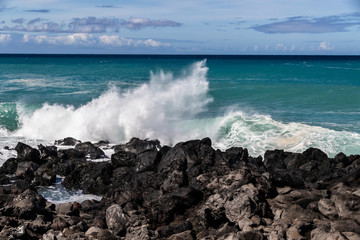 Fototapeta premium Wave breaking on the western Kona coast of Hawaii's Big Island near South Point. White sea spray thrown into the air; Deep blue-green Pacific ocean, and blue sky with clouds in the background. 