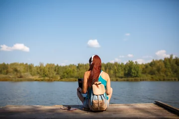 Foto op Canvas Photo on back of woman in headphones with backpack and laptop in hands sitting on river bank © Sergey