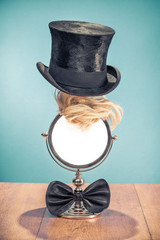 Retro silver mirror frame with wig hair, classic gentleman cylinder hat and black bow tie on aged oak wooden table front mint blue wall background. Barbershop concept. Vintage style filtered photo
