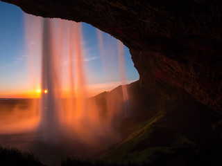 The waterfall Seljalandsfoss in the last golden light. One of the most famous waterfalls in Iceland. Shooted behind the waterfall at sunset. The sun's rays shine through the waterfall.