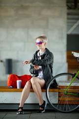 Photo of young woman in headphones with clock on hand sitting on bench next to green bicycle