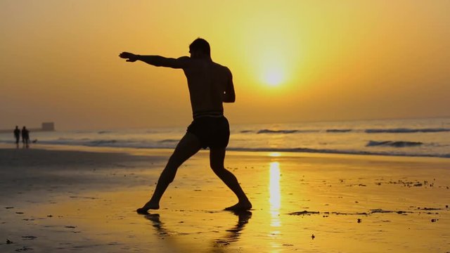 Muay thai fighter without t-shirt is doing shadow boxing on the beach during sunset