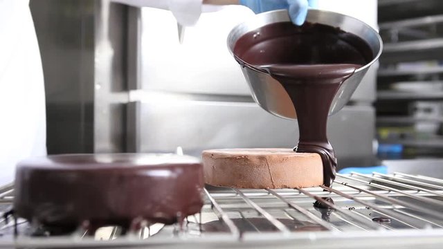 hands pastry chef prepares a cake, cover pouring chocolate icing, working on a stainless steel industrial kitchen work top