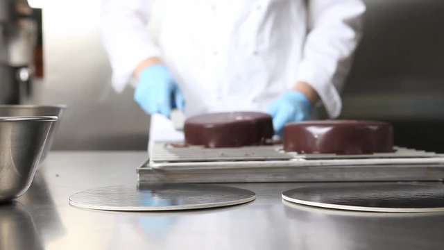 hands pastry chef prepares a cake, cover pouring chocolate icing, working on a stainless steel industrial kitchen work top