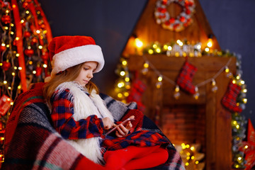 Fototapeta na wymiar Happy little girl in santa hat, plaid dress with fur playing with her smart phone while sitting on a chair by a fireplace in a cozy dark living room on Christmas eve