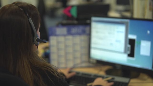 Unrecognizable female hotliner sitting at her desk in front of her computer screen in an open workspace, typing on her keyboard and talking to a customer through her phone headset