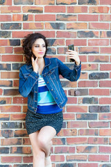 A young Caucasian woman with curly dark hair against red brick wall looking to a smartphone in her hands. Vertical