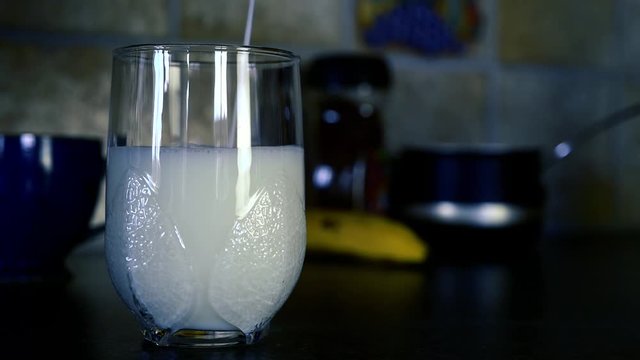 Milk is poured in an empty glass with fruits and kitchen equipment in background