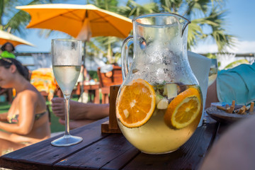 View of bar terrace and table with glass and jar of sangria in focus and background with people, parasol, sky and palm trees