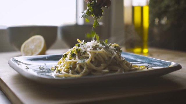 Pasta noodles on a square dish topped with grated cheese. Chopped parsley is dropped on top followed by a lemon squeezed over the food. Bright sunlight and a bottle of olive oil in back. Slow motion.