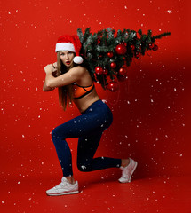 sexy Christmas fitness sport woman wearing santa hat holding xmas tree on her shoulders. snowflakes