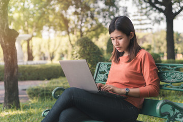 Happy young asian woman working with her laptop on a bench in the park outdoors on vacation time.