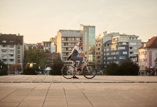 one young smiling man, 20-29 years old,  wearing hipster suit, smart casual, sitting on old city bike. city buildings panorama behind in background. full lenght shot.