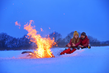 Happy loving couple sitting at a fire in the snow among the fields in the winter against the...
