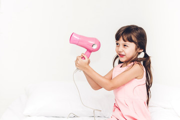 Smiling little girl dries the hair dryer sitting at home on the bed